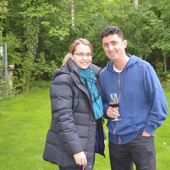 Ein Mann und eine Frau stehen auf einem Rasen und lächeln für ein Foto. Der Mann hält ein Weinglas in der Hand, und sie sind von Bäumen umgeben.