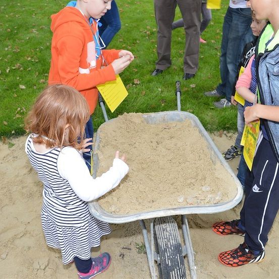 Ein junges Mädchen schiebt einen mit Sand gefüllten Schubkarren, während ein Junge zusieht. Andere Kinder und Erwachsene stehen um sie herum auf einem Rasen.