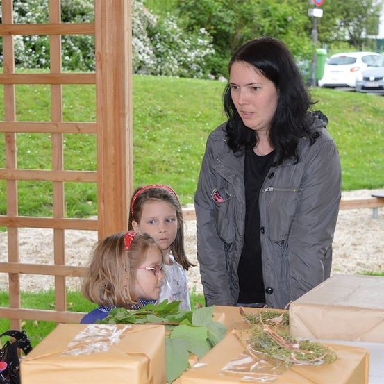 Eine Frau steht mit zwei jungen Mädchen vor einem Tisch mit verpackten Geschenken in einem Garten mit einem Holzgitter und einer Rasenfläche.