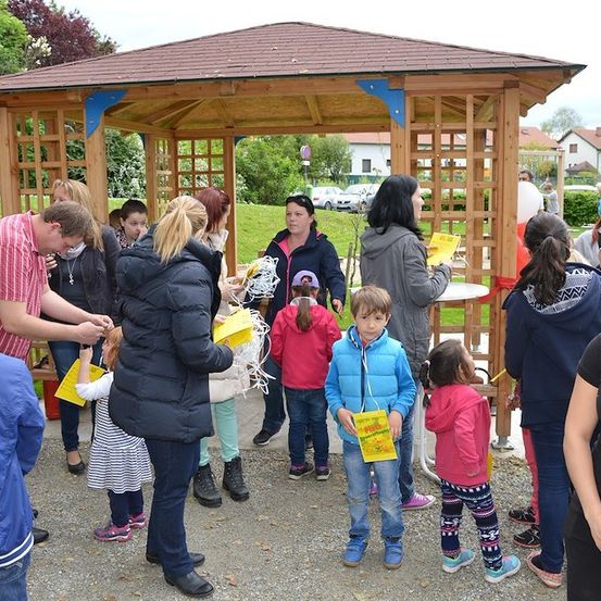 Eine Gruppe von Menschen, einschließlich Kindern, versammelt sich um einen hölzernen Pavillon, einige halten gelbe Papiere. Eine Frau in einer grauen Kapuzenjacke hält ein gelbes Papier und schaut darauf. Der Pavillon ist von Bäumen und Gras umgeben.