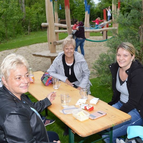 Drei Frauen sitzen an einem Picknicktisch in einem Park und lächeln. Dahinter ist eine hölzerne Spielstruktur zu sehen, auf der Kinder spielen. Im Hintergrund befinden sich üppige Grünflächen und ein klarer blauer Himmel.
