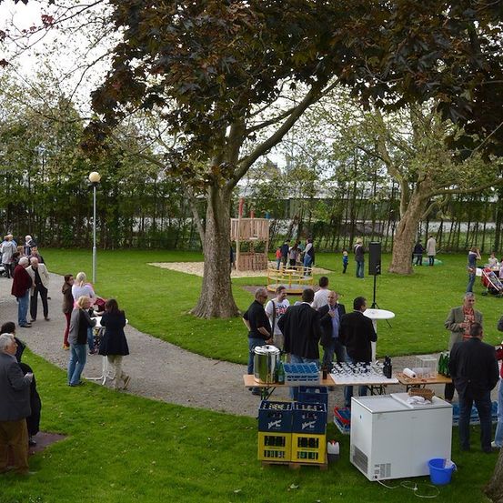 Ein Park mit Menschen, die sich unterhalten, einem Tisch mit Getränken und einem Spielplatz im Hintergrund.