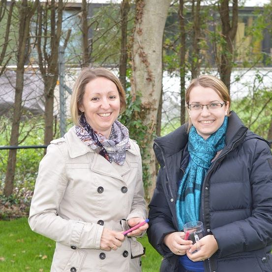 Zwei Frauen stehen in einem Park, lächeln und halten Stifte. Eine trägt einen beigen Mantel und eine Schürze, die andere einen blauen Schal und eine schwarze Jacke. Sie sind von Bäumen und einem Zaun umgeben.
