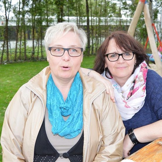 Zwei Frauen sitzen auf einer Bank in einem Park. Die Frau auf der linken Seite hat kurze weiße Haare und trägt einen blauen Schal. Die Frau auf der rechten Seite hat einen Blumenschal und trägt ein blaues Hemd. Dahinter befindet sich ein Spielplatz mit einer Rutsche.