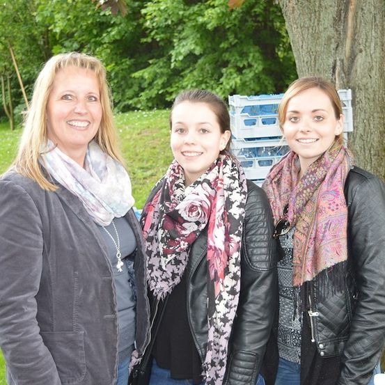 Drei Frauen stehen zusammen in einem Park, lächeln und posieren für ein Foto. Die Frau auf der linken Seite trägt eine graue Jacke und einen Schal. Die Frau in der Mitte trägt eine schwarze Lederjacke und einen Blumenschal. Die Frau auf der rechten Seite trägt eine schwarze Lederjacke und einen gemusterten Schal. Hinter ihnen steht ein Baumstamm und einige Lagerboxen.