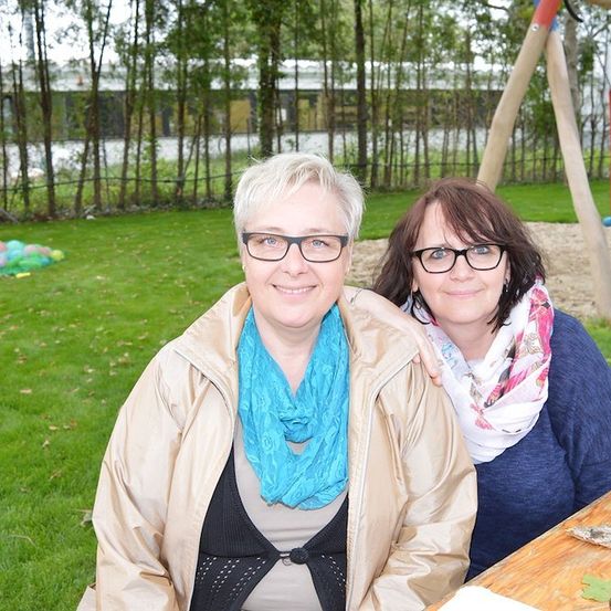 Zwei Frauen mit Brille sitzen auf einer Bank in einem Park mit einem Spielplatz im Hintergrund.
