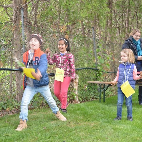 Eine Gruppe von Kindern ist im Freien in einem Park, ein Kind hält ein gelbes Objekt und scheint es zu werfen, während andere gelbe Papiere halten. Eine Frau steht hinter ihnen.