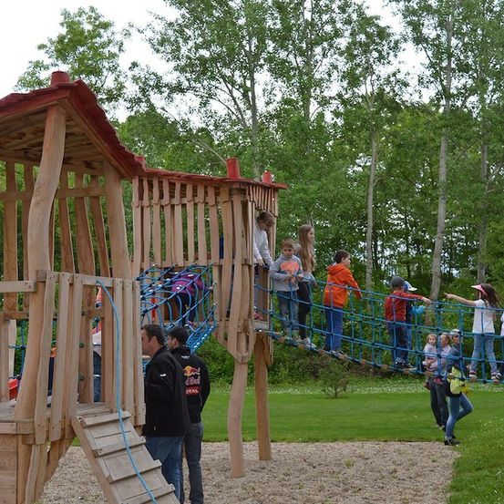 Eine Gruppe von Kindern und Erwachsenen spielt auf einer hölzernen Spielstruktur mit einer Seilbrücke und Kletternetz. Der Spielplatz ist von Bäumen und Gras umgeben.