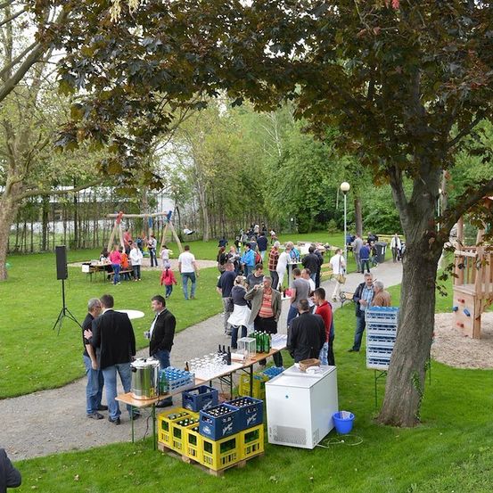 Ein Gemeindetreffen in einem Park mit Menschen, die sich unterhalten, einem Tisch mit Getränken und einem Spielplatz im Hintergrund.