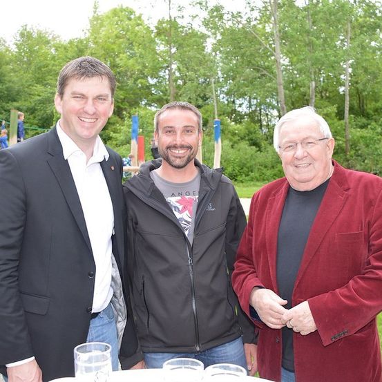 Drei Männer stehen lächelnd für ein Foto in einem Park. Einer trägt einen schwarzen Anzug, ein anderer eine graue Kapuzenjacke und der dritte einen roten Mantel. Hinter ihnen sind ein Spielplatz und üppige Grünflächen zu sehen.