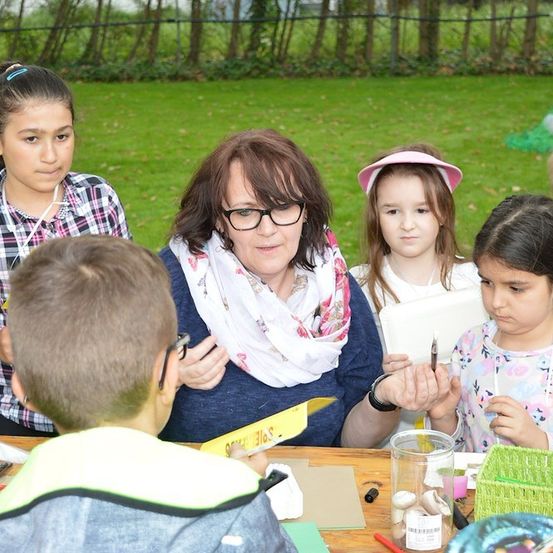Eine Gruppe von Kindern und einer erwachsenen Frau versammelt sich um einen Tisch in einem Park und ist in einer Aktivität vertieft. Die Frau hält einen Stift und Papier, und die Kinder schauen zu ihr. Auf dem Tisch befinden sich verschiedene Gegenstände, darunter ein Glas und ein Korb. Im Hintergrund ist eine Rasenfläche mit Bäumen und einem Zaun zu sehen.