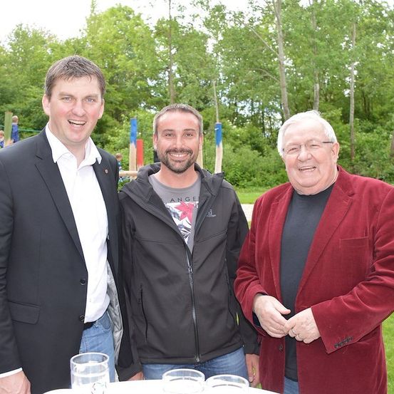 Drei Männer stehen lächelnd für ein Foto in einem Park. Der Mann links trägt einen schwarzen Anzug, der Mann in der Mitte eine schwarze Jacke und der Mann rechts eine rote Samtjacke. Hinter ihnen ist ein Spielplatz mit Rutschen und Bäumen zu sehen.