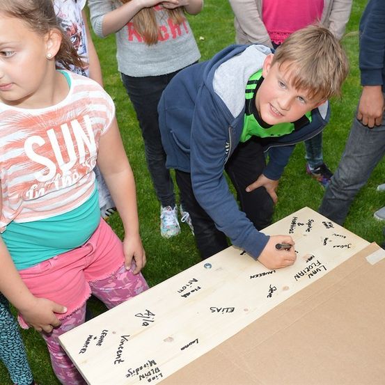 Ein junger Junge schreibt auf ein Pappschild mit vielen Namen und Nachrichten, während ein Mädchen neben ihm steht. Sie sind draußen auf einem Grasfeld mit anderen Leuten im Hintergrund.