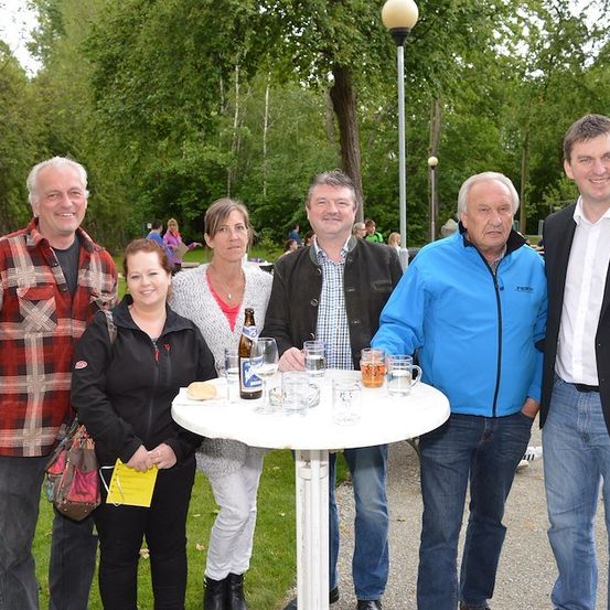 Eine Gruppe von Menschen steht um einen Tisch mit Getränken und Essen herum, lächelt und posiert für ein Foto im Park.