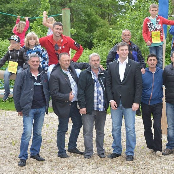 Eine Gruppe von Erwachsenen und Kindern posiert für ein Foto in einem Park mit einem Spielplatz im Hintergrund. Die Erwachsenen stehen vor einer Holzstruktur.