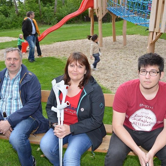 Drei Personen sitzen auf einer Parkbank. Eine Frau in der Mitte hält eine Krücke. Hinter ihnen läuft ein Kind auf dem Spielplatz, und ein Mann steht in der Nähe einer Rutsche.
