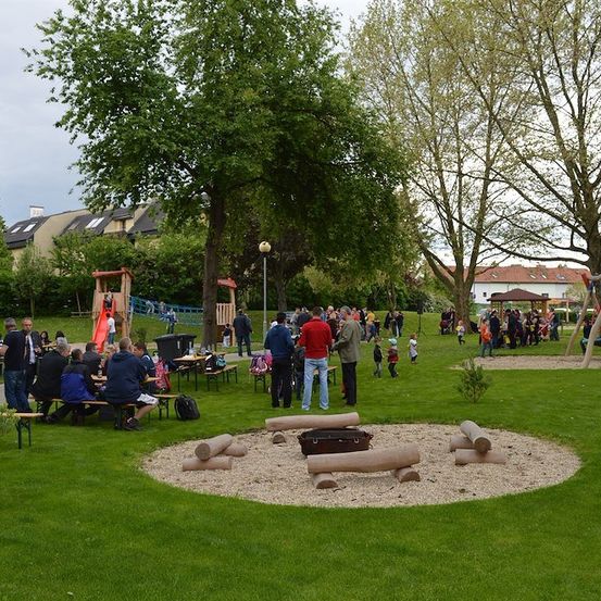 Ein Park mit Spielplatz, Picknicktischen und einem Feuerkorb. Menschen sind versammelt, einige sitzen und andere stehen. Bäume und Gebäude im Hintergrund.