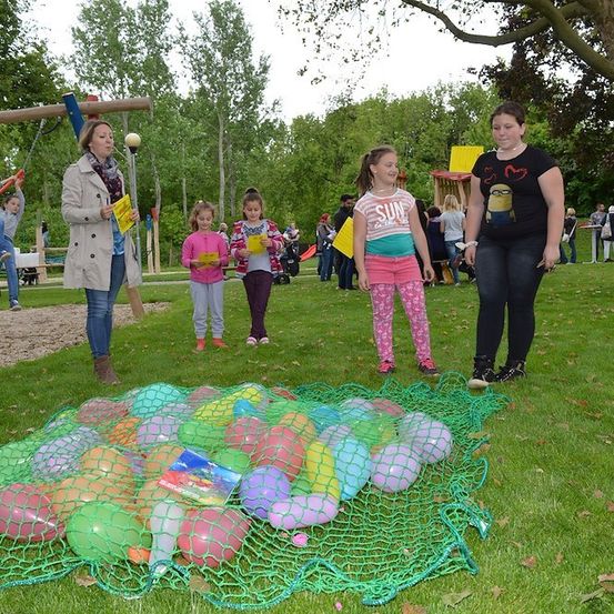 Eine Gruppe von Kindern und Erwachsenen versammelt sich in einem Park, eine Frau hält ein gelbes Papier. In der Nähe liegt ein Netz voller bunter Luftballons auf dem Gras.