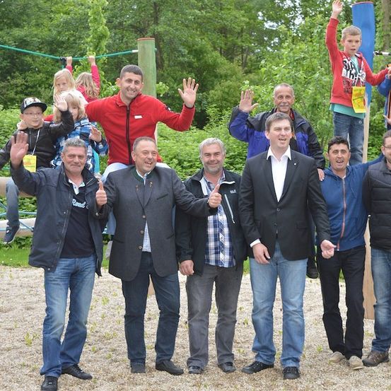 Eine Gruppe von Erwachsenen und Kindern posiert für ein Foto in einem Park, wobei einige auf einer Holzstruktur stehen und andere winken. Bäume und Grünflächen sind im Hintergrund zu sehen.