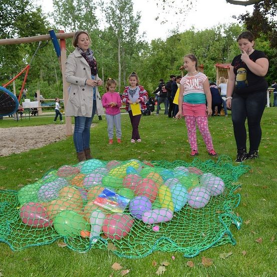 Eine Gruppe von Kindern und Erwachsenen versammelt sich um ein großes Netz, das mit bunten Luftballons gefüllt ist, auf einem Rasen. Die Erwachsenen stehen, während die Kinder die Ballons betrachten. Im Hintergrund befinden sich Bäume und ein Spielplatz.