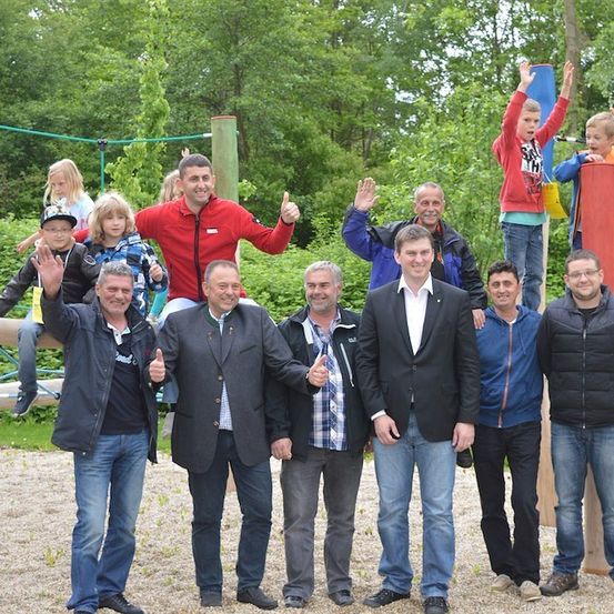 Eine Gruppe von Menschen, einschließlich Erwachsener und Kindern, steht zusammen in einem Park mit Spielplatzgeräten im Hintergrund. Sie alle lächeln und geben Daumen hoch.