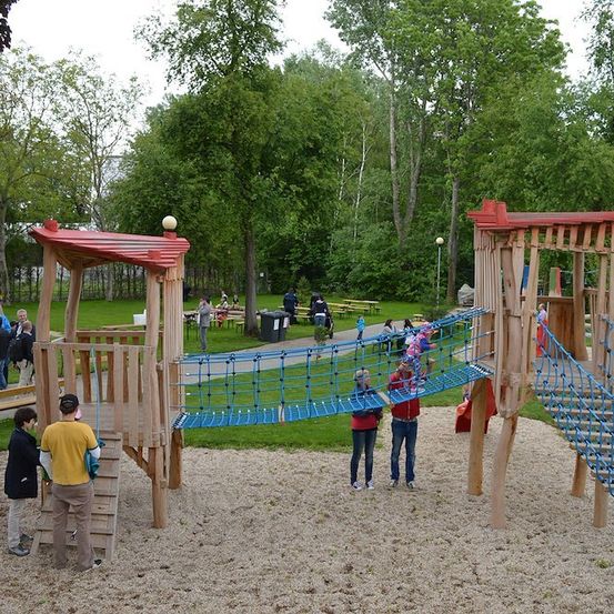 Ein Spielplatz mit einer hölzernen Kletterstruktur und einer Netzbrücke, umgeben von einem Rasenbereich mit Bäumen und Menschen.