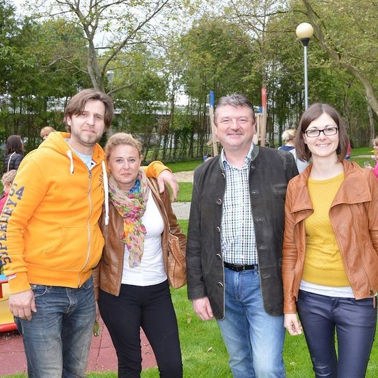 Vier Personen stehen in einem Park, lächeln und posieren für ein Foto. Sie sind lässig gekleidet, und im Hintergrund ist ein Spielplatz zu sehen.