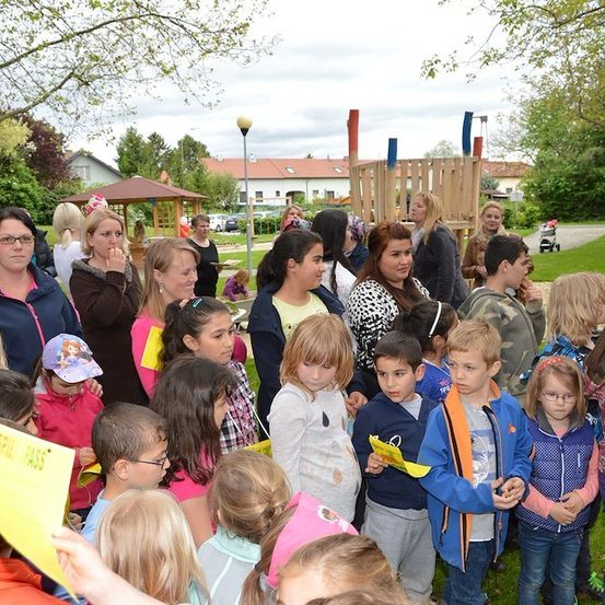 Eine Gruppe von Kindern und Erwachsenen versammelt sich in einem Park, einige halten gelbe Papiere. Der Park hat ein Spielplatz, Bäume und Häuser im Hintergrund.