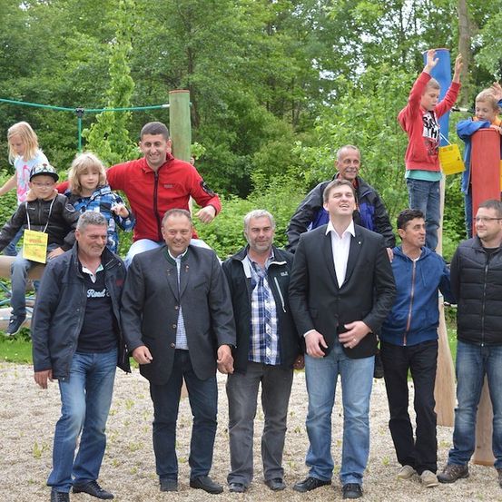 Eine Gruppe von Erwachsenen und Kindern posiert für ein Foto in einem Park mit einem Spielplatz im Hintergrund. Die Erwachsenen stehen vor einer Holzstruktur, während die Kinder darauf klettern.