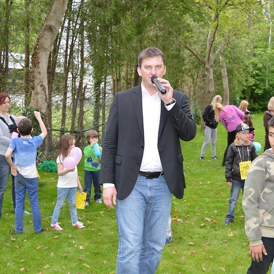 Ein Mann in einem Anzug spricht in ein Mikrofon, während er in einem Park mit Kindern und Erwachsenen steht. Einige Kinder halten Luftballons und andere gehen herum.