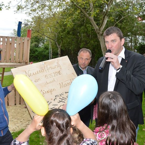 Ein Mann in einem Anzug spricht in ein Mikrofon und hält einen gelben Luftballon, hinter ihm ein Holzschild mit der Aufschrift 'Neverisfrung am 13.05.2016 durch Milan Marsc'. Zwei Mädchen halten blaue und gelbe Luftballons hoch.