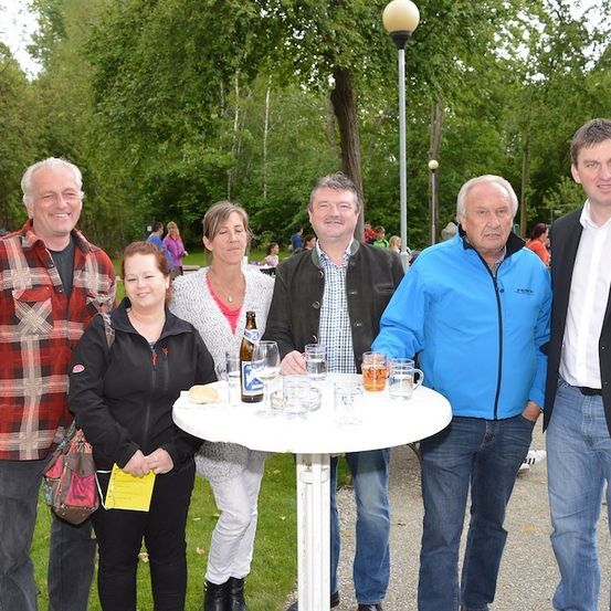 Eine Gruppe von Menschen steht um einen Tisch in einem Park, lächelt und posiert für ein Foto. Auf dem Tisch sind Gläser und eine Flasche. Hinter ihnen sind Bäume und eine Straßenlaterne.