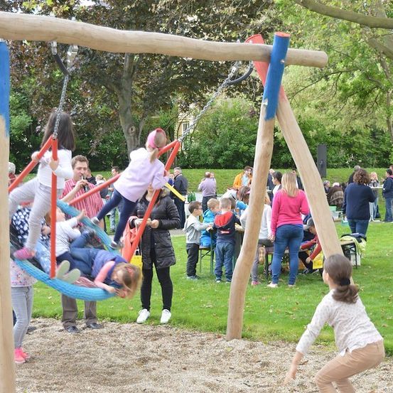 Mehrere Kinder spielen auf Schaukeln in einem Park, während Erwachsene beaufsichtigen und andere im Hintergrund plaudern.