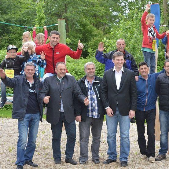 Eine Gruppe von Männern und Kindern posiert für ein Foto in einem Park mit einem Spielplatz im Hintergrund.