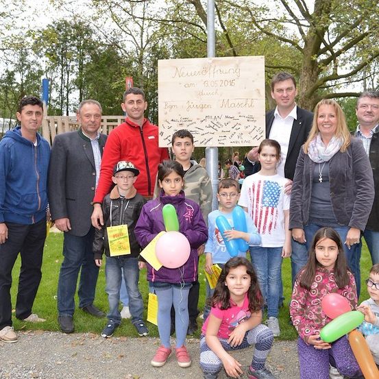 Eine Gruppe von Menschen, einschließlich Erwachsener und Kindern, steht in einem Park, lächelt und posiert für ein Foto. Ein Schild im Hintergrund lautet 'Neueröffnung am 03.05.2016'. Die Kinder halten Luftballons.