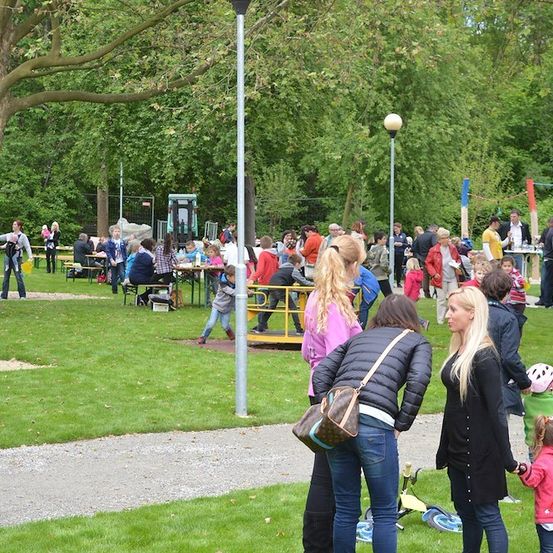 Ein Park mit Menschen, die um Tische und einen Spielplatz herum versammelt sind, einige tragen Jacken und haben Taschen, unter einem klaren Himmel.
