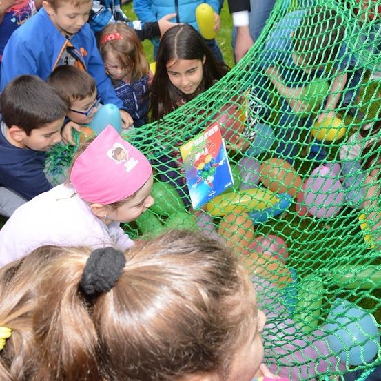 Eine Gruppe von Kindern spielt in einem Netz voller bunter Luftballons. Einige halten Luftballons, während andere ein Buch betrachten.