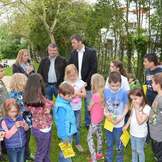 Eine Gruppe von Kindern und Erwachsenen versammelt sich in einem Park, einige halten gelbe Papiere, während zwei Männer in Anzügen in der Mitte stehen, einer hält ein Mikrofon.