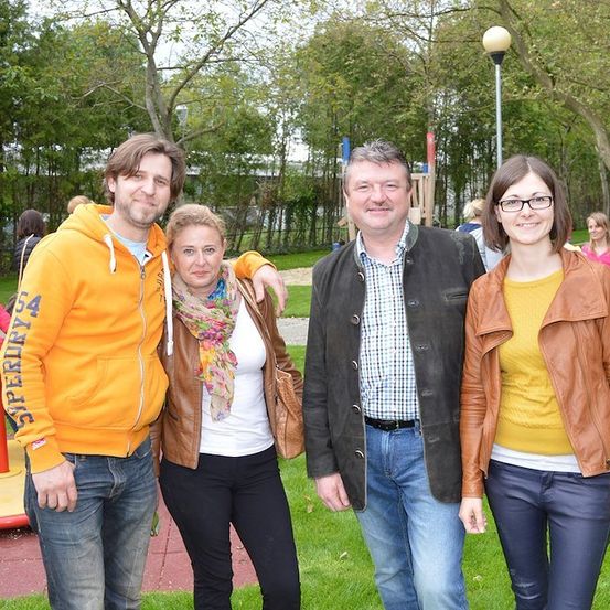 Vier Menschen stehen in einem Park und lächeln für ein Foto. Ein Mann in einer orangefarbenen Kapuzenjacke steht links, eine Frau mit einem Schal in der Mitte und ein Mann in einer Jacke rechts. Eine Frau in einer Lederjacke steht ganz rechts. Hinter ihnen befindet sich ein Spielplatz mit einer Rutsche und einer Bank.