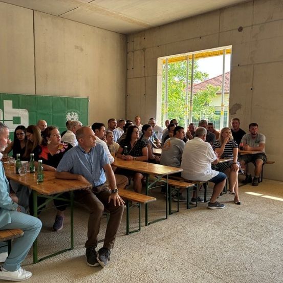 Eine Gruppe von Menschen sitzt um Tische in einem Raum mit einem großen Fenster und einem Banner an der Wand.