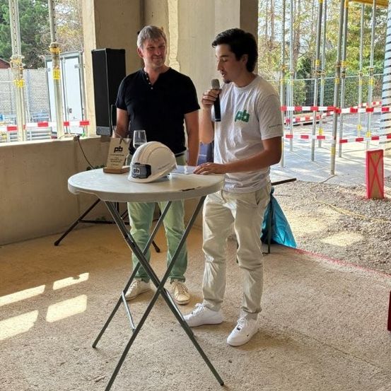Zwei Männer stehen an einem Tisch, einer hält ein Mikrofon und spricht, während der andere einen Schutzhelm hält und zuhört. Dahinter befindet sich eine Baustelle mit Gerüsten.