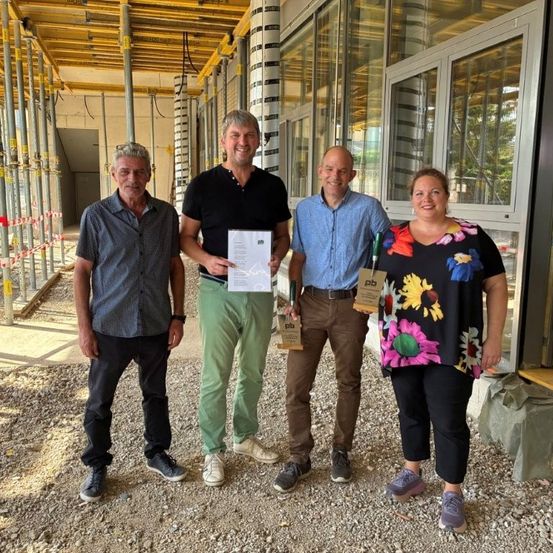 Vier Personen stehen vor einem im Bau befindlichen Gebäude und lächeln für ein Foto. Ein Mann hält ein Zertifikat und ein anderer ein Trophäe.