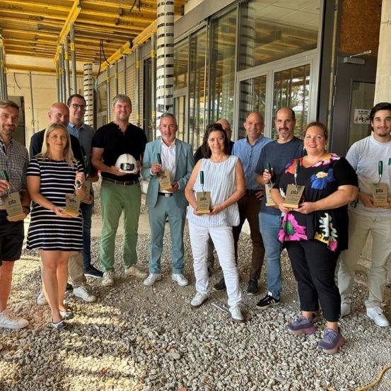 Eine Gruppe von Menschen, wahrscheinlich Kollegen, steht vor einem im Bau befindlichen Gebäude, hält Auszeichnungen in der Hand und lächelt für ein Foto.