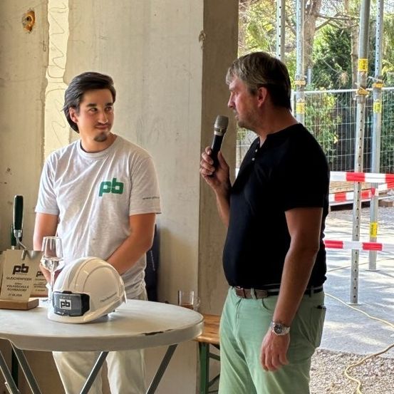 Zwei Männer stehen vor einem Tisch mit einem Mikrofon, einer in einem weißen Shirt mit grünem Logo, der andere in einem schwarzen Shirt. Dahinter ein Baustelle mit einem weißen Helm auf dem Tisch.