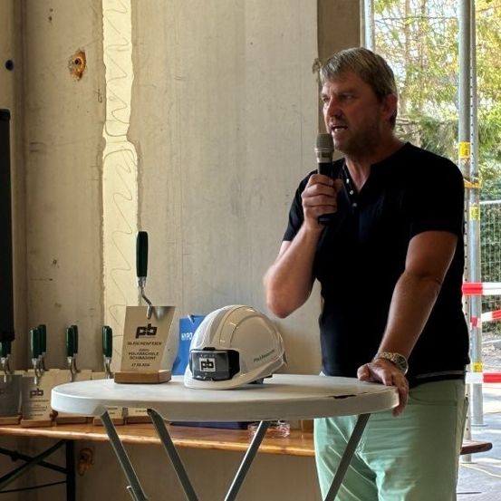 Ein Mann steht an einem Tisch und hält ein Mikrofon in der Hand, mit einem Bauhelm und Werkzeug auf dem Tisch. Er ist in einem schwarzen Shirt und grüner Hose gekleidet. Hinter ihm befindet sich eine Wand mit einem Fenster und ein sichtbarer Baustelle.