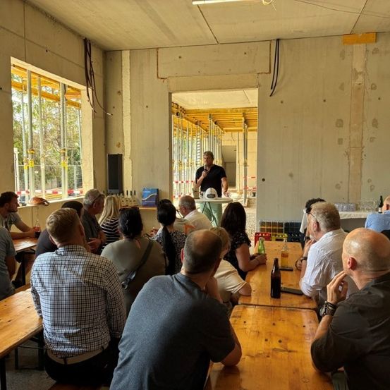 Eine Gruppe von Menschen sitzt um Holztische in einem Raum im Bau herum, mit einem Mann, der vor ihnen steht, möglicherweise eine Präsentation hält.