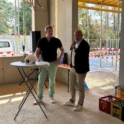 Zwei Männer stehen auf einer Baustelle, einer hält ein Mikrofon und spricht, der andere steht neben einem Tisch mit einem Helm darauf. Hinter ihnen sind Gerüste und eine Wand zu sehen.
