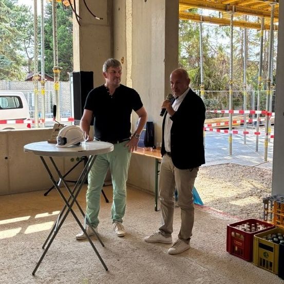 Zwei Männer stehen auf einer Baustelle, einer hält ein Mikrofon und spricht, der andere steht neben einem Tisch mit einem Helm darauf. Hinter ihnen sind Gerüste und eine Wand zu sehen.