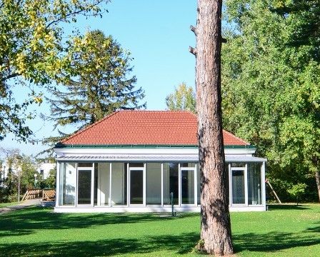 Ein kleines weißes Gebäude mit rotem Dach und großen Fenstern steht in einem Park, umgeben von Bäumen und Gras.