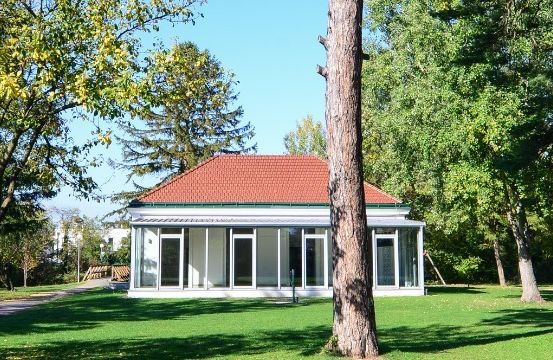 Ein kleines weißes Gebäude mit rotem Dach und großen Fenstern steht in einem Park, umgeben von Bäumen und Gras.